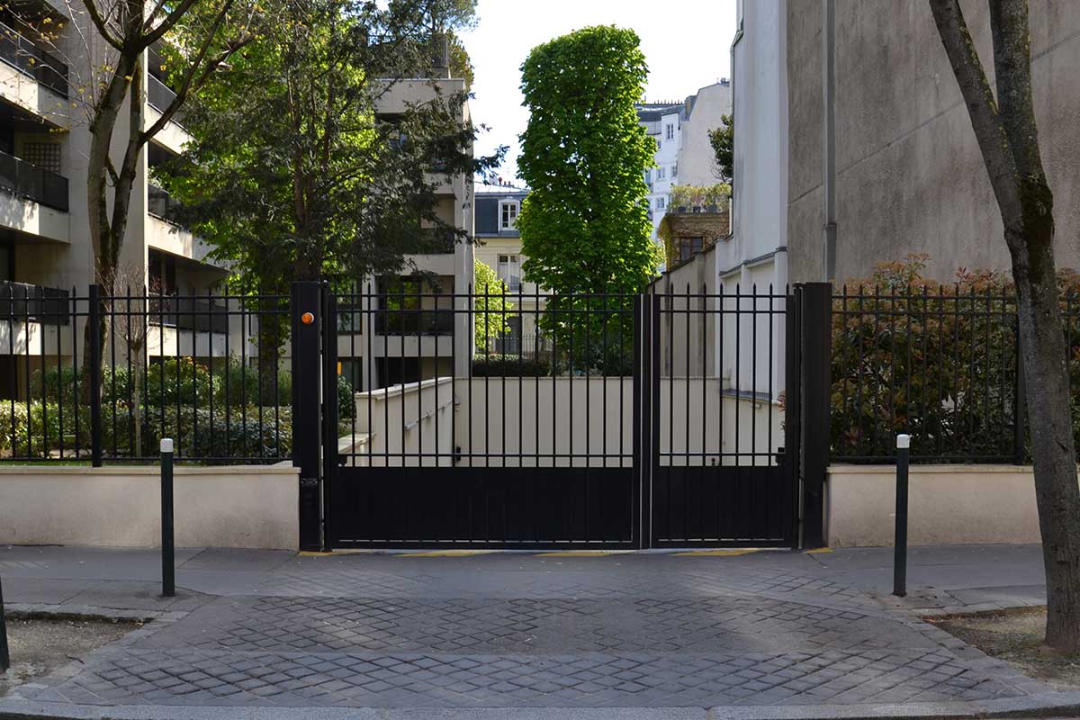 Closed black metal gate blocking access to a light-colored residential courtyard with trees and buildings.