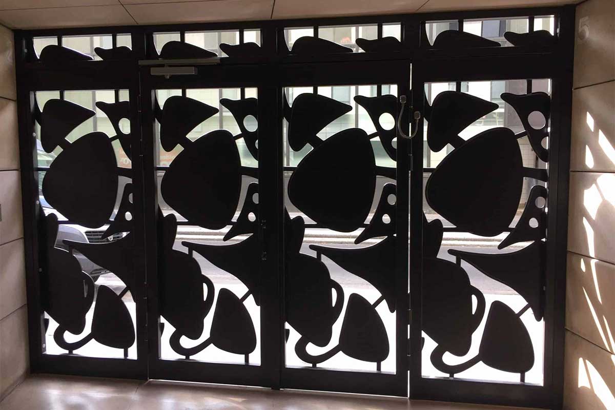 SERFA-Porte-de-hall-en-acier-sur-mesure-sécurité-et-design-pour-une-entrée-dimmeuble-2 | SERFA 1982 Entrance door with four glass panels covered by decorative black metal screens featuring large abstract leaf shapes.
